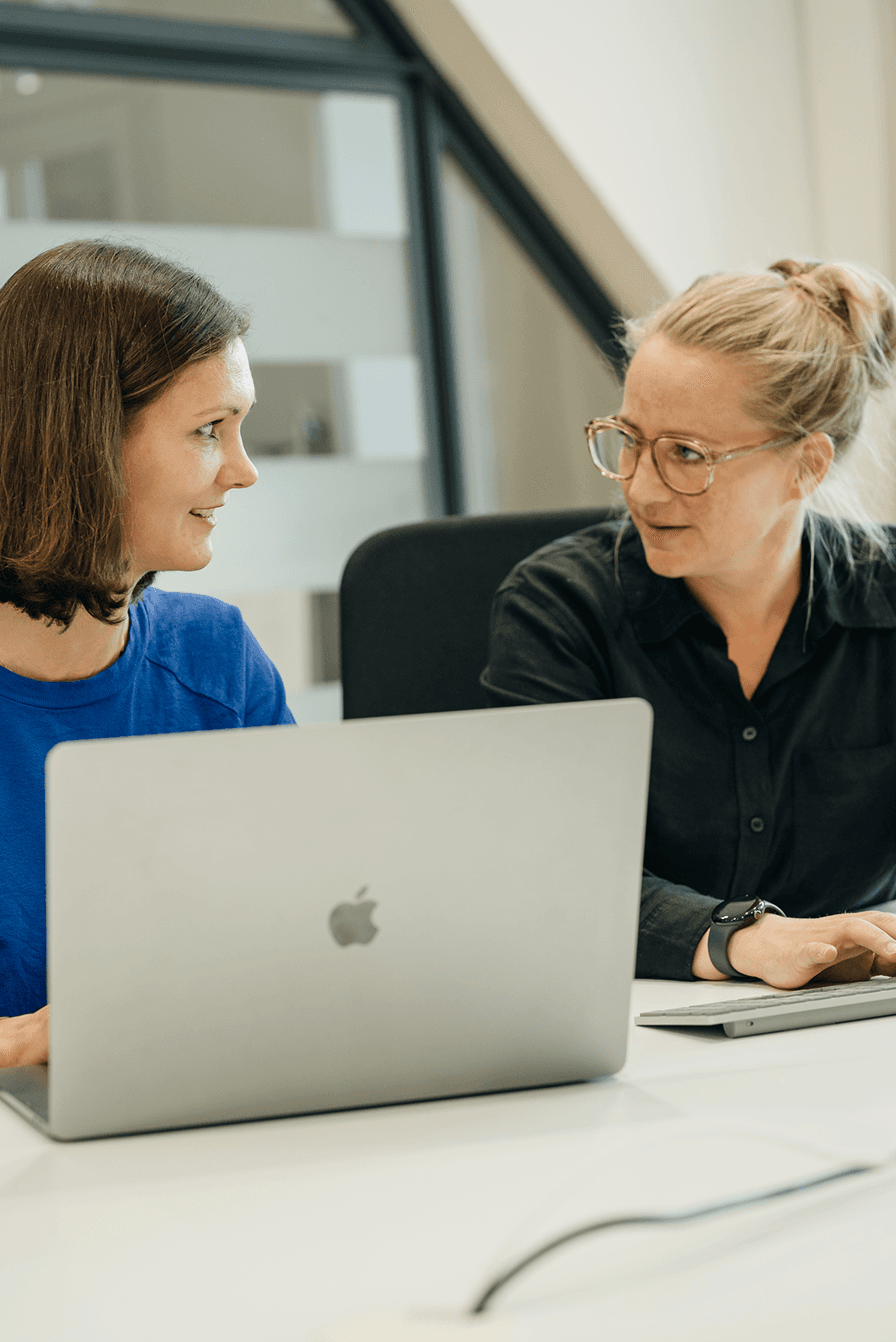 Zwei Frauen sitzen an einem Tisch in einem hellen Büro und unterhalten sich. Auf dem Tisch steht ein Laptop.