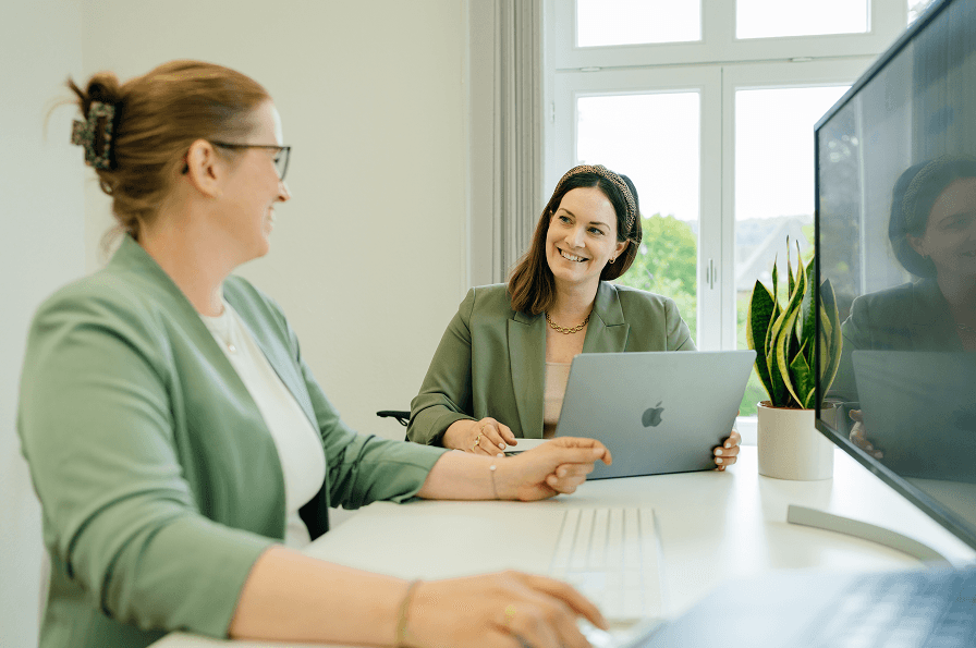 Zwei Frauen in grünen Blazern sitzen an einem Schreibtisch mit Laptop und Monitor, lächeln und unterhalten sich in einem hellen Büro.