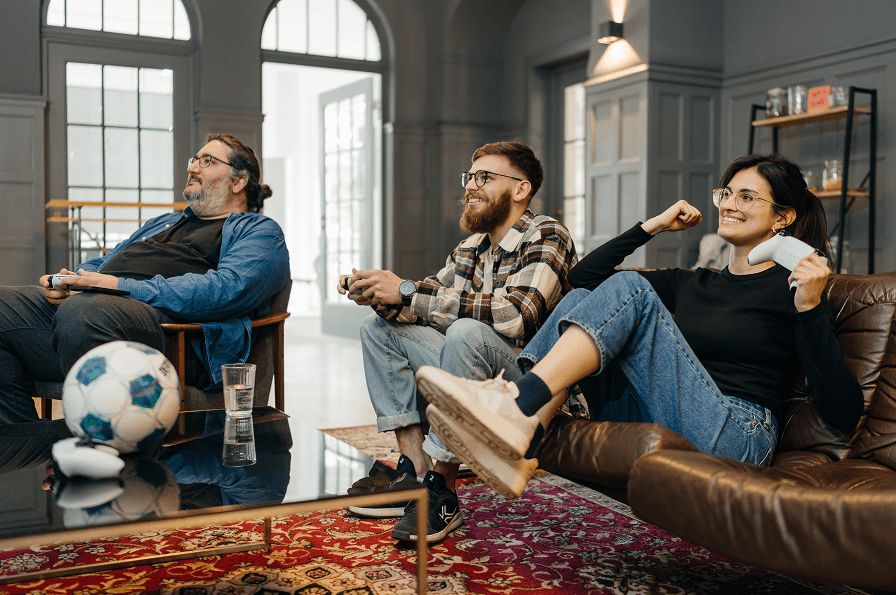 Drei Personen sitzen auf einem Sofa in einem hellen Raum und spielen Videospiele. Alle halten Controller in der Hand, die Frau auf der rechten Seite schaut triumphierend und auf dem Tisch liegt ein Fußball.
