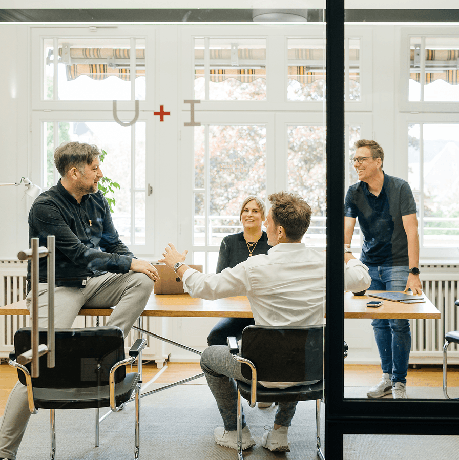 Vier Personen führen eine Diskussion an einem Tisch in einem hellen Büro mit großen Fenstern. An der Glastür ist ein Schild mit der Aufschrift "u and i" zu sehen.