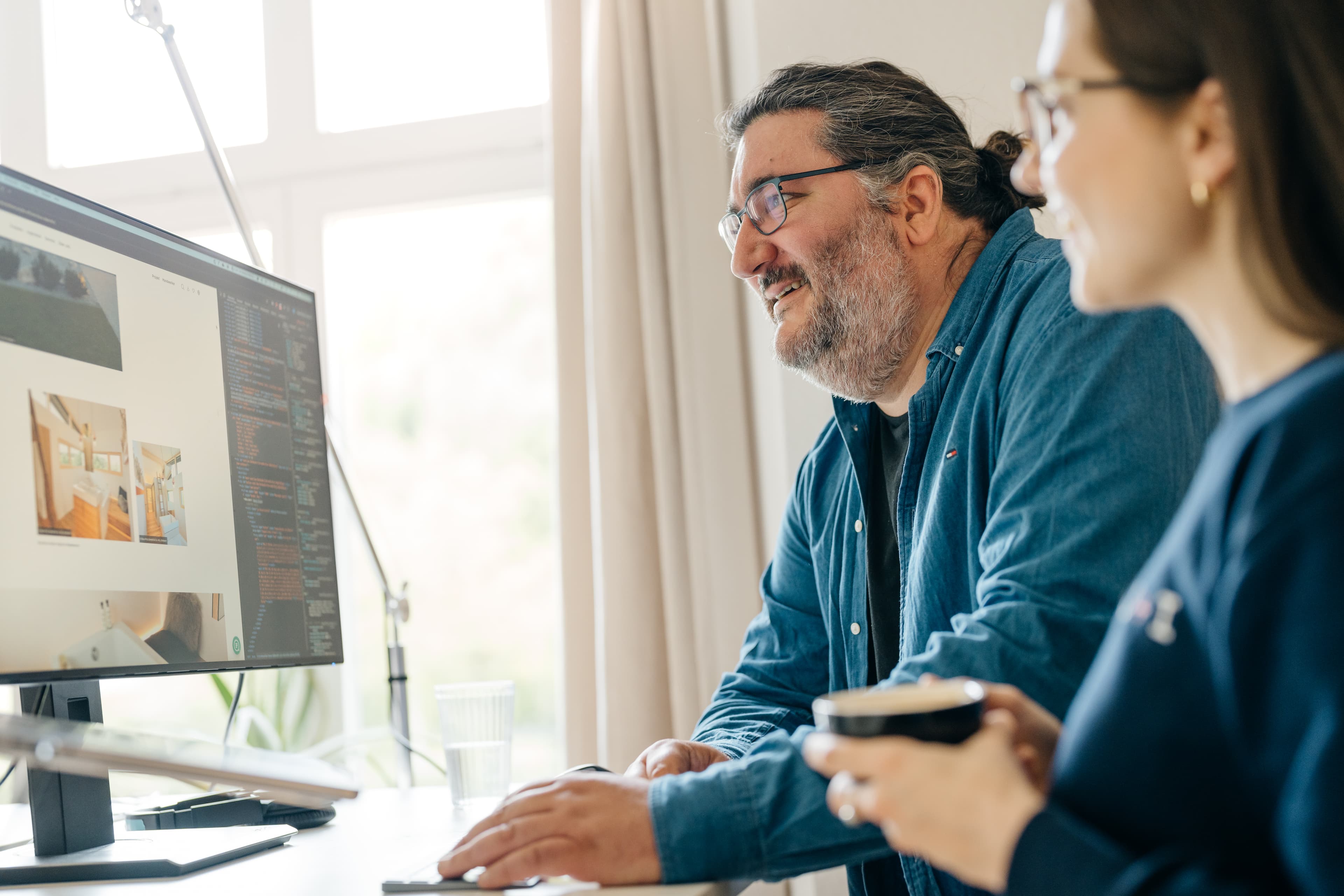 Zwei Personen sitzen an einem Schreibtisch und schauen auf einen Computerbildschirm mit einer Website. Eine Person hält eine Tasse. Der Raum ist hell.