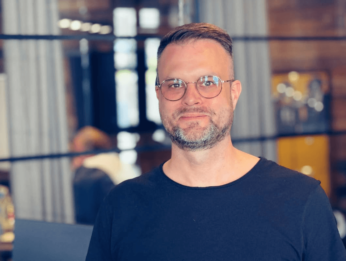 Ein Mann mit Brille und gepflegtem Bart, der ein schwarzes Hemd trägt, steht in einem modernen Büro mit Holzwänden und Glastrennwänden.