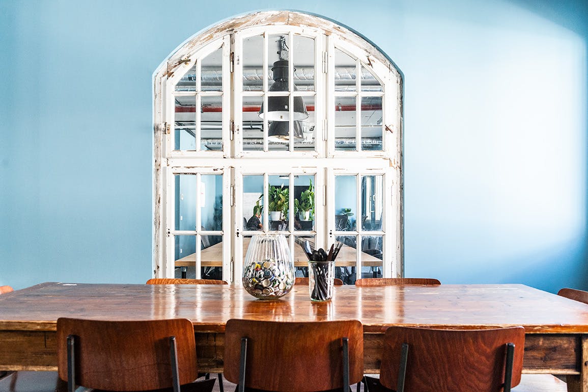 Ein Blick auf den schönen alten Holztisch, der in unserem Bielefelder Büro in der Küche als Esstisch dient. Herum stehen Vintage-Holzstühle. Die Wand im Hintergrund ist in hellem Türkis-Blau gestrichen, durch ein großes Bogenfenster fällt der Blick auf einen der offenen Büroräume. u+i interact