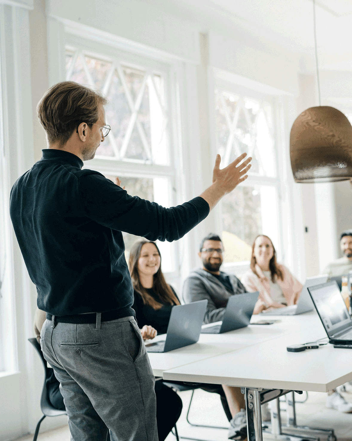 Ein Mann steht und gestikuliert, während er zu einer Gruppe von Menschen spricht, die mit Laptops an einem Tisch in einem hellen Besprechungsraum sitzen.