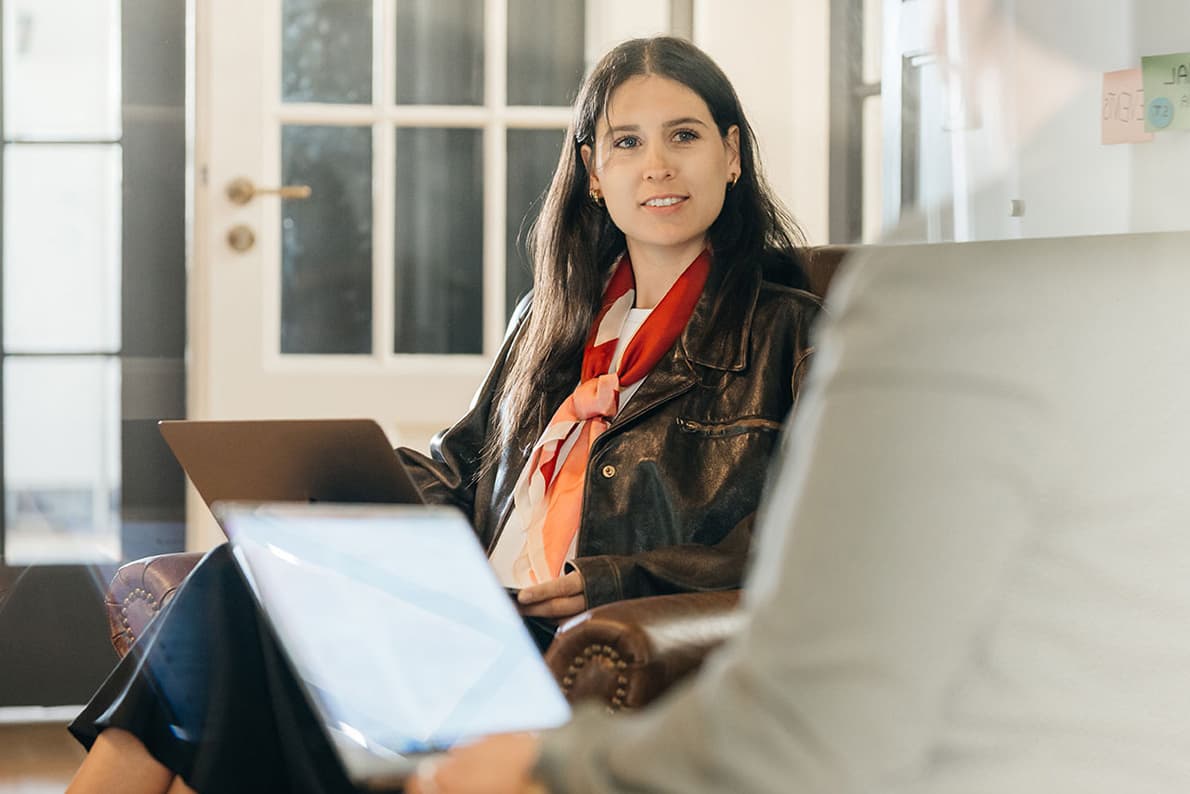 Eine Frau, die mit einem Laptop auf einem Stuhl sitzt und eine Lederjacke sowie einen roten Schal trägt, unterhält sich mit einer unscharfen Person im Vordergrund.
