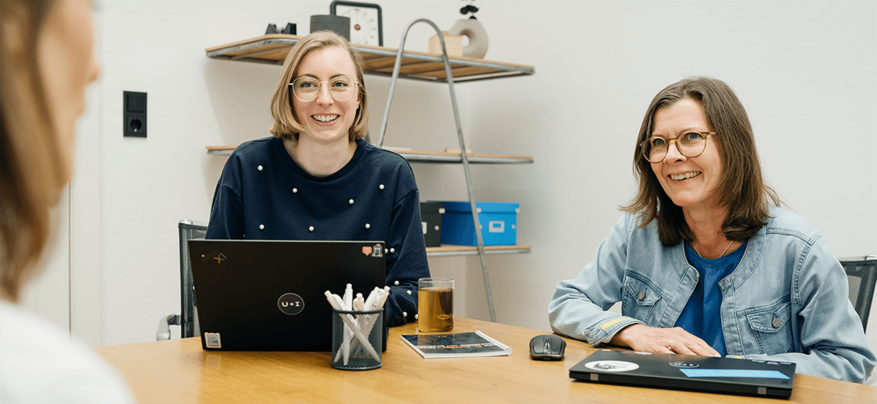 Zwei Frauen sitzen an einem Holztisch mit ihren Laptops, lächeln und unterhalten sich mit jemandem, der nicht im Bild zu sehen ist.