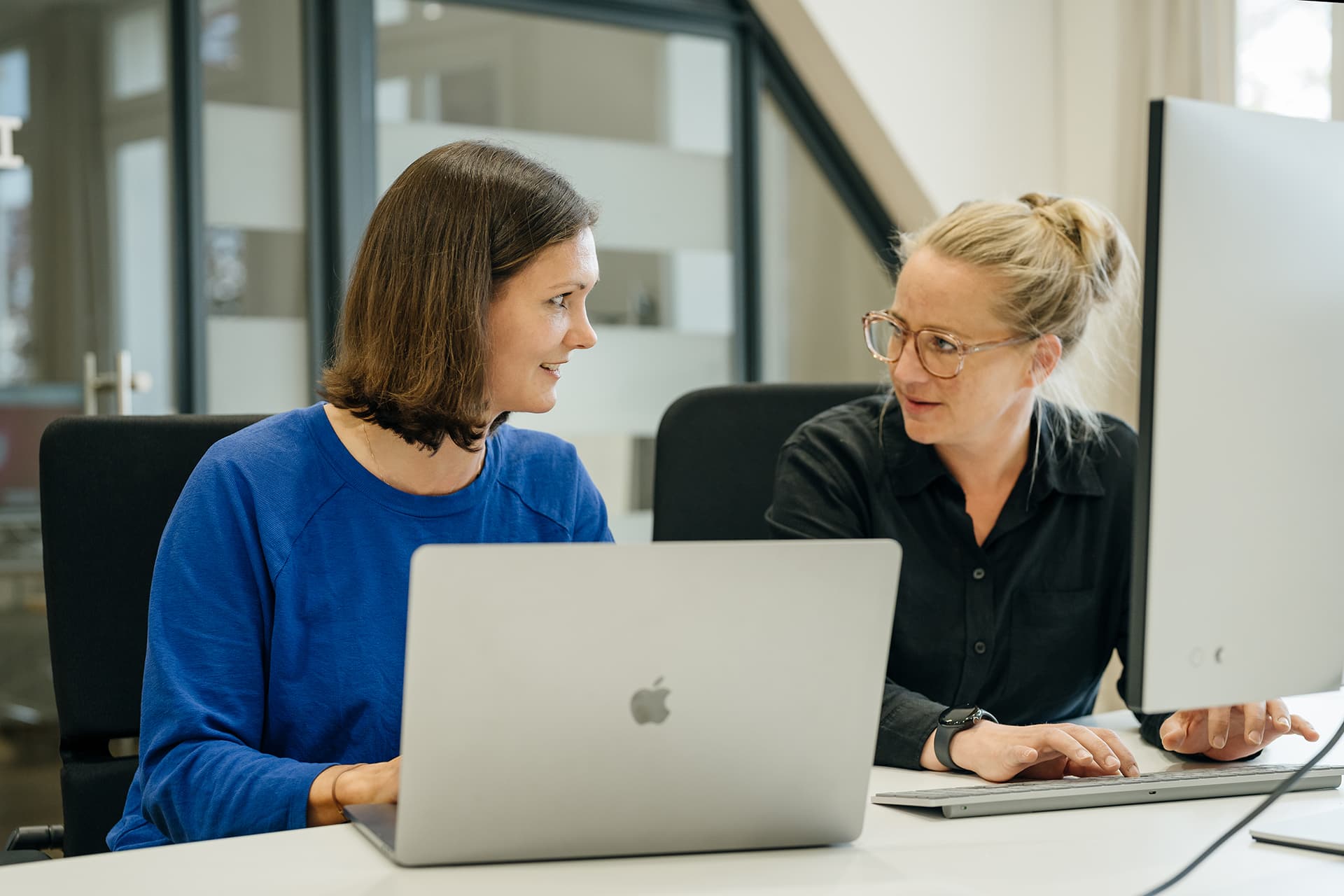 Zwei Frauen sitzen an einem Schreibtisch und unterhalten sich; die eine arbeitet an einem Laptop, die andere an einem Desktop-Computer.