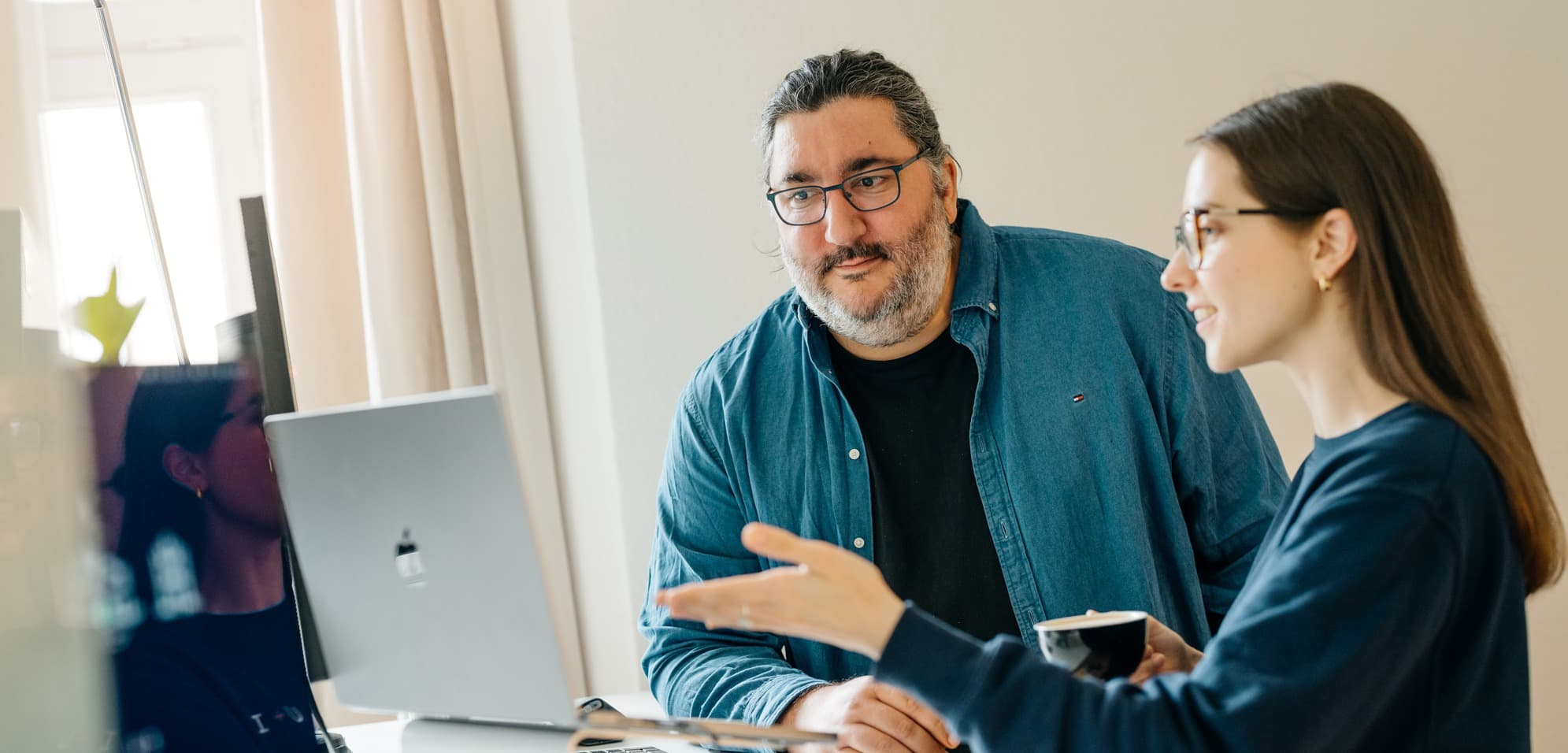 Eine Frau gestikuliert, während sie sich mit einem Mann unterhält. Sie stehen an einem Schreibtisch mit Laptops und einer Tasse in einem gut beleuchteten Raum eines modernen Büros.