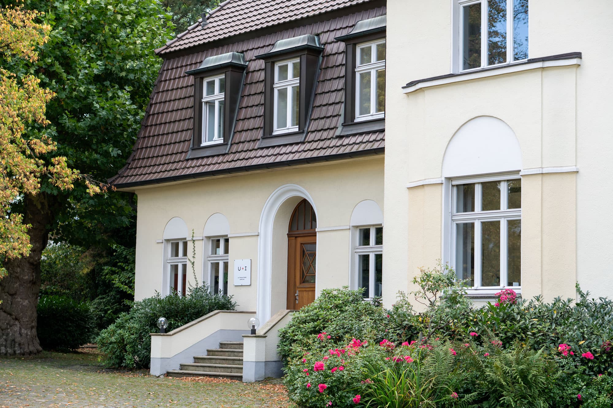 Außenansicht einer hellen Villa mit braunem Dach und weißen Fenstern. Vor dem Gebäude führen Treppen mit Geländern zum Eingangsbereich. Links neben der Tür befindet sich ein Schild mit der Aufschrift „U+I“. Der Eingangsbereich ist von gepflegten Sträuchern und blühenden Rosen umgeben.