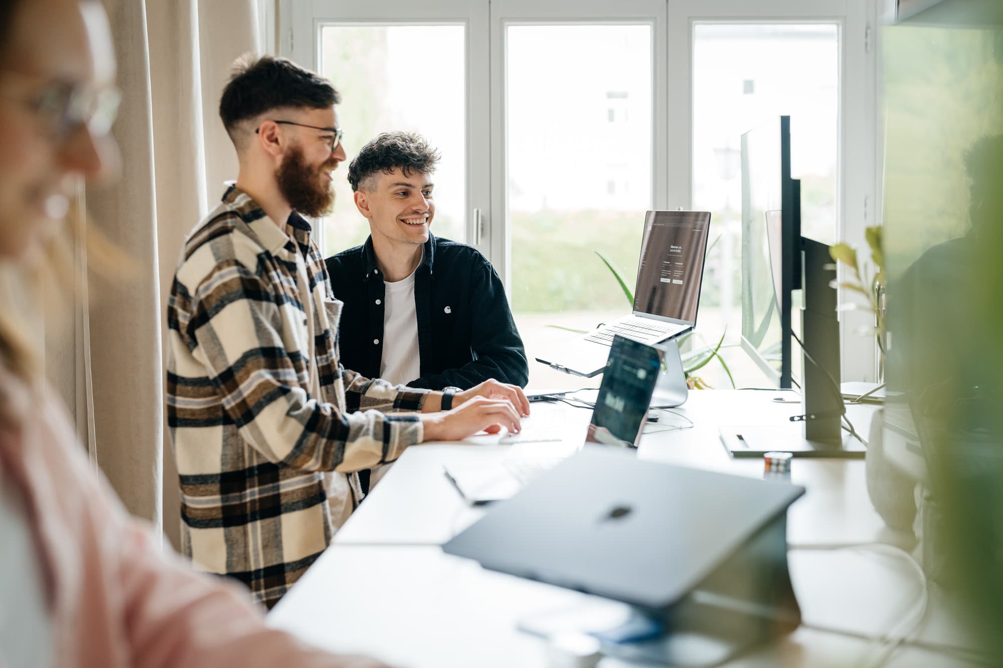 Zwei Kollegen stehen lachend nebeneinander an einem Steharbeitsplatz in einem hellen Büro. Auf dem Laptop-Ständer vor ihnen ist eine Website zu sehen. Die Atmosphäre wirkt locker und kollegial.