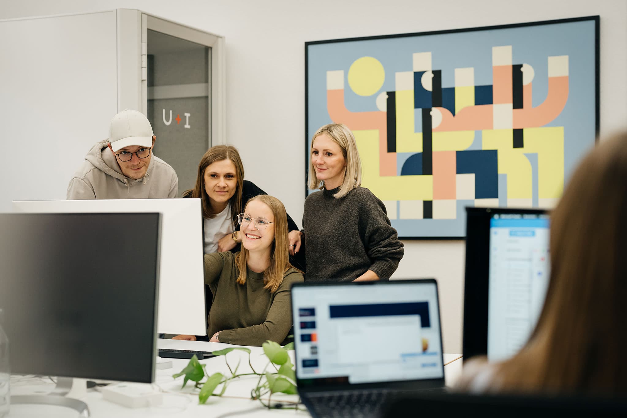 Vier Personen in einem Büro stehen um einen Schreibtisch herum und schauen auf einen Computerbildschirm. An der Wand hinter ihnen hängt ein farbenfrohes abstraktes Gemälde.