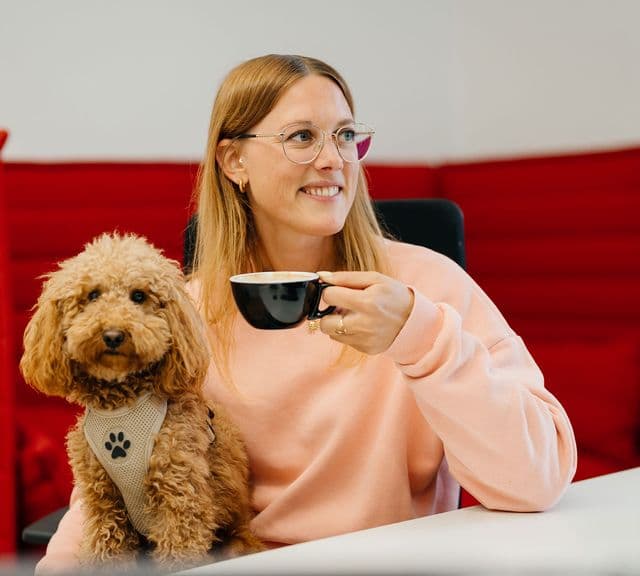 Lächelnde Frau mit Brille hält eine Kaffeetasse in der Hand, während sie an einem Schreibtisch sitzt, neben ihr ein flauschiger brauner Hund mit Geschirr. Im Hintergrund eine rote Wand.