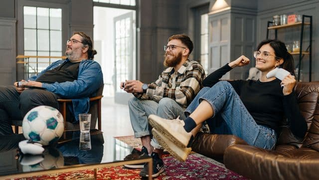 Drei Personen sitzen auf einem Sofa in einem Wohnzimmer und spielen begeistert Videospiele, während auf dem Tisch vor ihnen ein Fußball und ein Glas stehen.