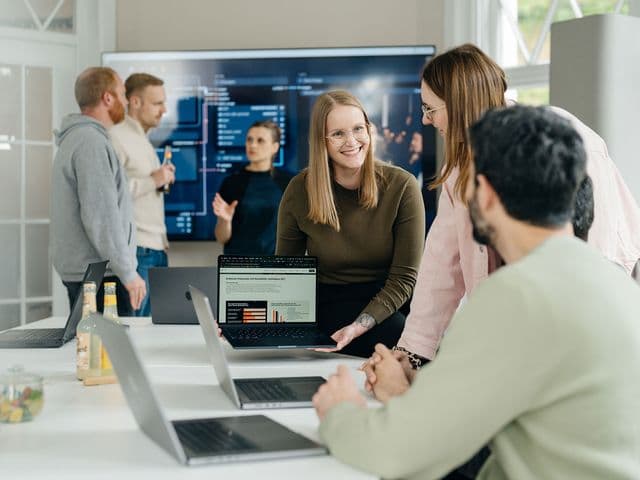 Eine Gruppe von Menschen in einer modernen Büroumgebung betrachtet und diskutiert Informationen auf einem Laptop mit Diagrammen, während andere an einem großen Bildschirm arbeiten.