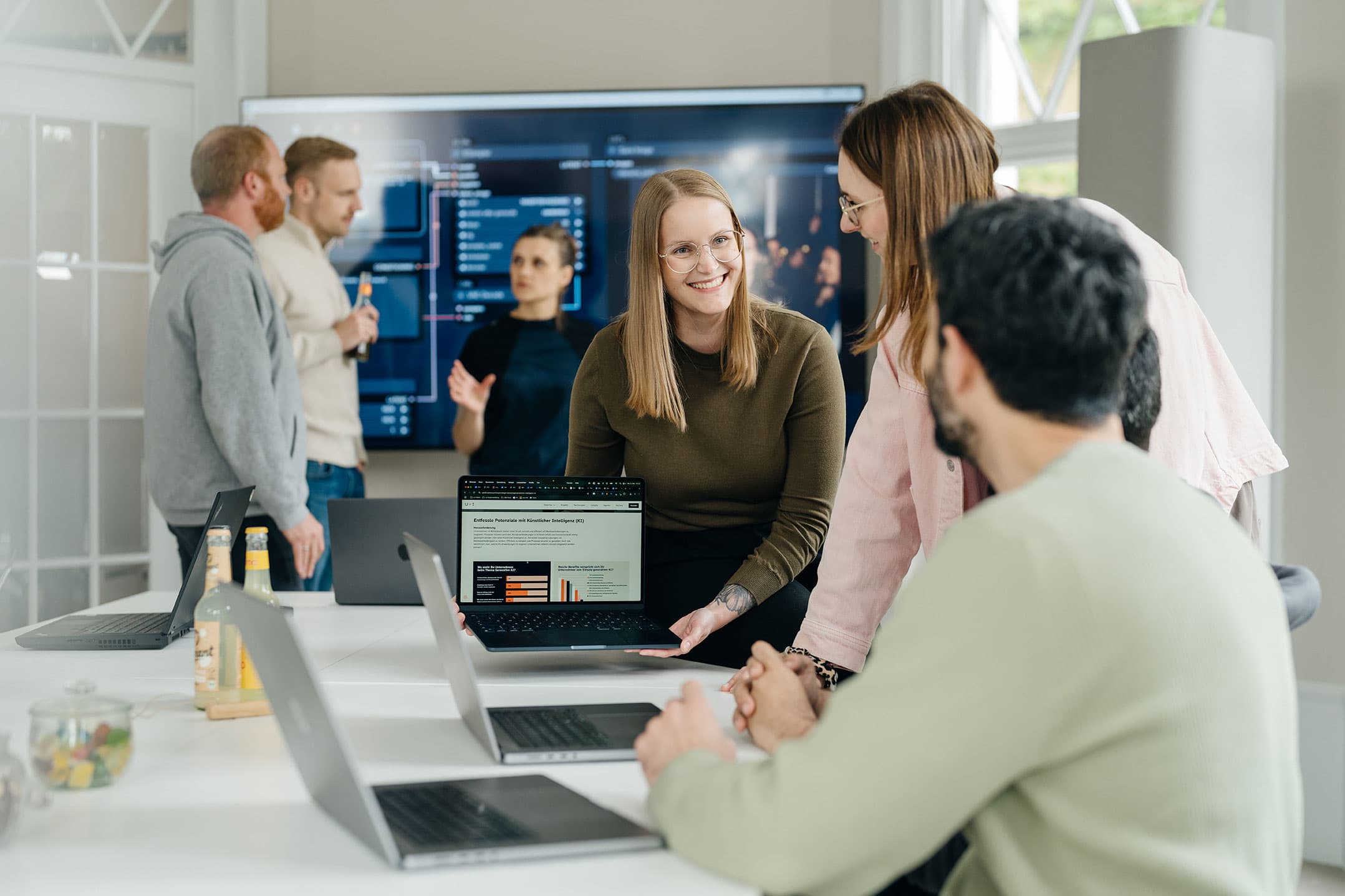 Eine Gruppe von Menschen in einer modernen Büroumgebung betrachtet und diskutiert Informationen auf einem Laptop mit Diagrammen, während andere an einem großen Bildschirm arbeiten.