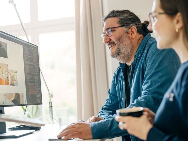 Zwei Personen lächeln und arbeiten am Computer, während sie Bilder von Innenarchitektur betrachten. Eine Person hält eine Kaffeetasse in der Hand. Heller Raum mit großem Fenster.