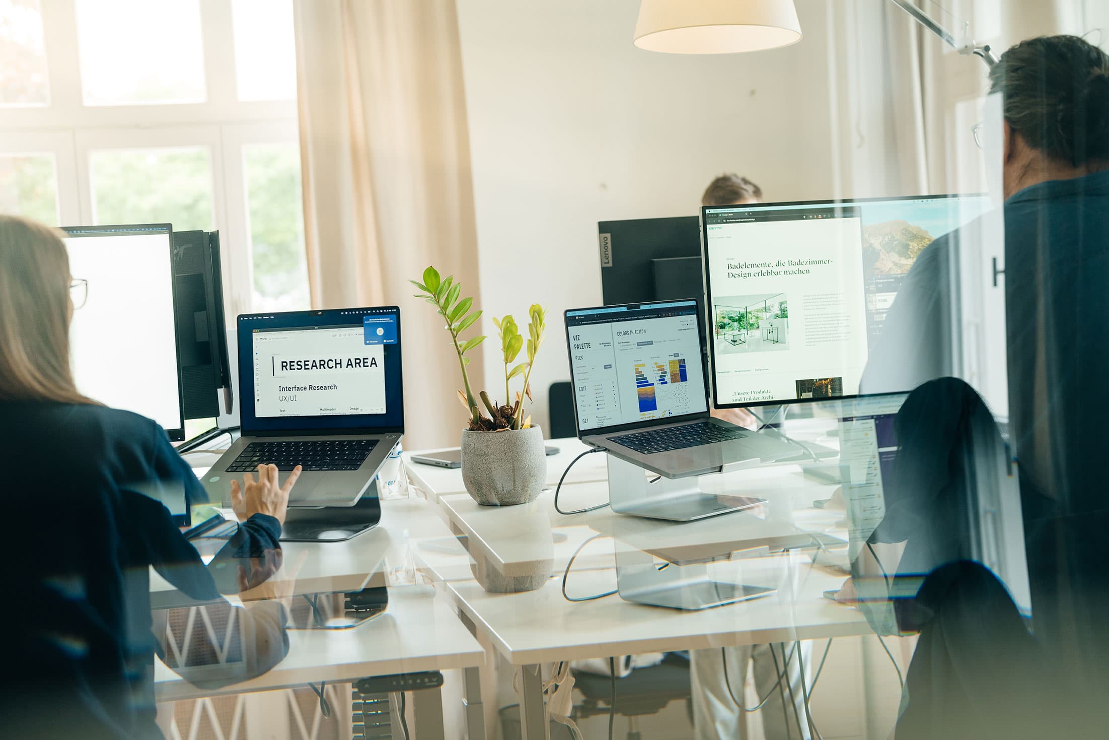 Blick durch eine Glasscheibe in ein helles Büro, in dem mehrere Personen an Schreibtischen arbeiten. Auf den Bildschirmen sind UX/UI-Arbeitsflächen zu sehen – darunter ein „Research Area"-Board und eine Farbpaletten-Ansicht. Im Hintergrund ist eine fertige Website zu erkennen.