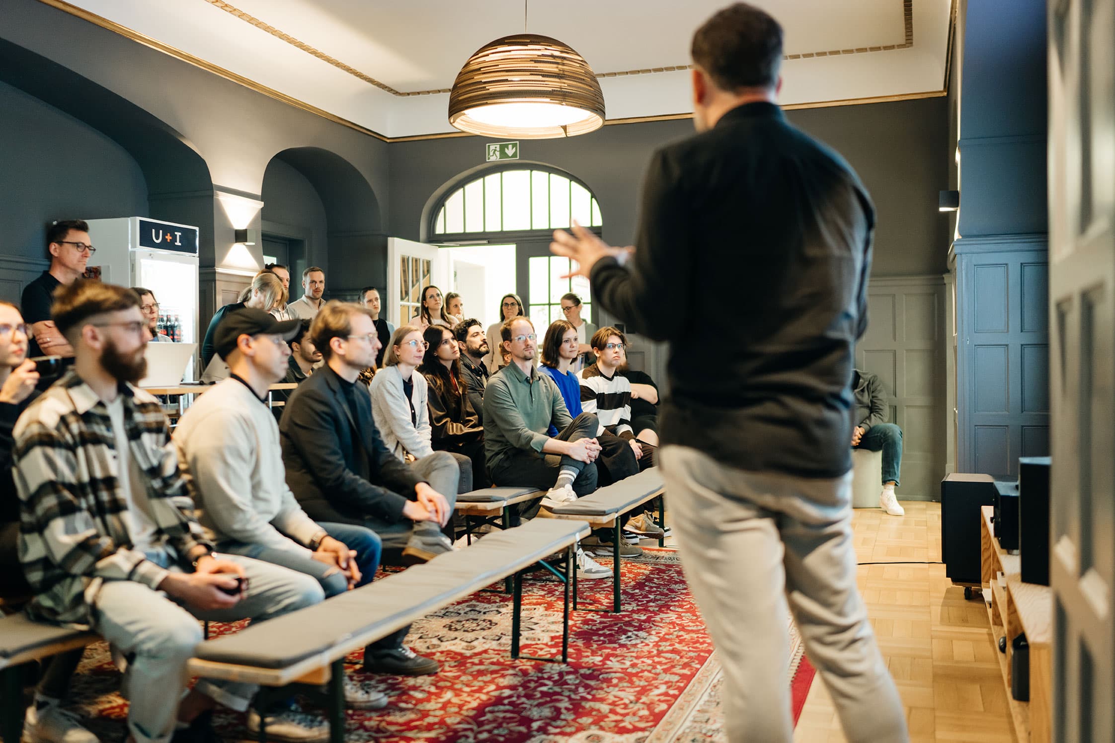 Ein Redner spricht zu einem aufmerksamen Publikum, das auf Bänken in einem gut beleuchteten Raum mit einer großen Pendelleuchte und einem dekorativen Teppich sitzt.