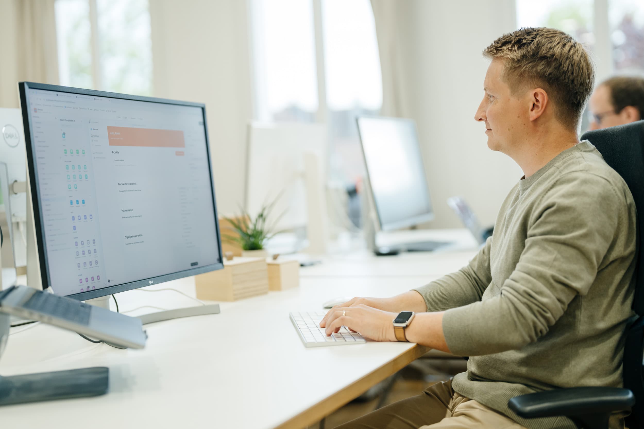 Ein Mann sitzt an einem hellen Schreibtisch in einem modernen Büro und arbeitet konzentriert an einem großen LG-Monitor, auf dem eine digitale Projektmanagement-Oberfläche zu sehen ist. Im Hintergrund ist ein weiterer Arbeitsplatz mit Bildschirm erkennbar.