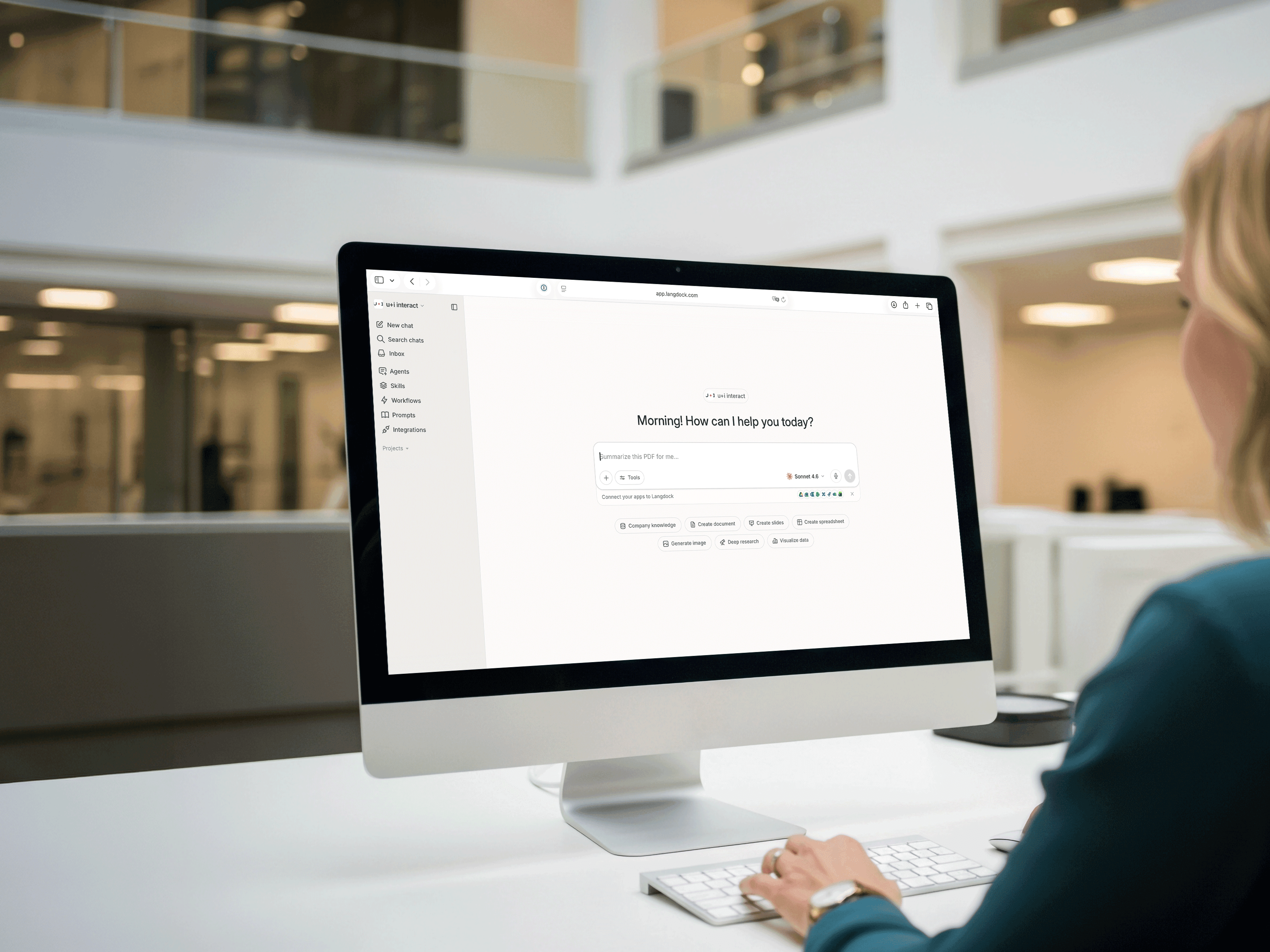 Eine Person in einem Büro sitzt an einem Schreibtisch vor einem verschwommenen Bürohintergrund und nutzt einen Computer, auf dem die Benutzeroberfläche des Langdock-Assistenten angezeigt wird.