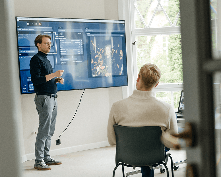 Ein Mann hält in einem hellen Raum eine Präsentation, auf einer großen Leinwand sind eine Software-Oberfläche und ein Foto zu sehen, während ein anderer Mann aufmerksam zuhört.