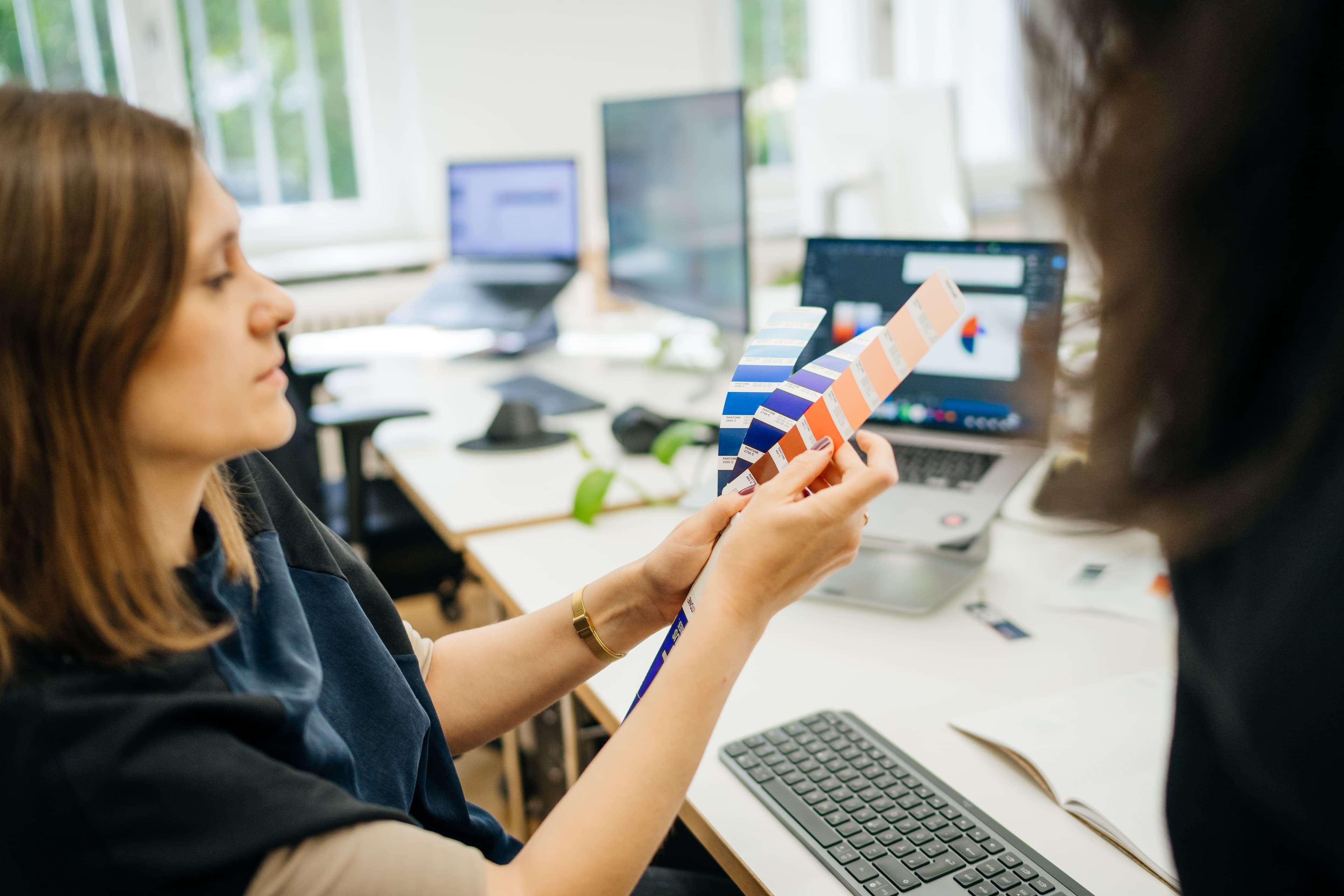 Eine Frau hält ein Farbmuster in der Hand und vergleicht Farbtöne, im Hintergrund sind Computer zu sehen.