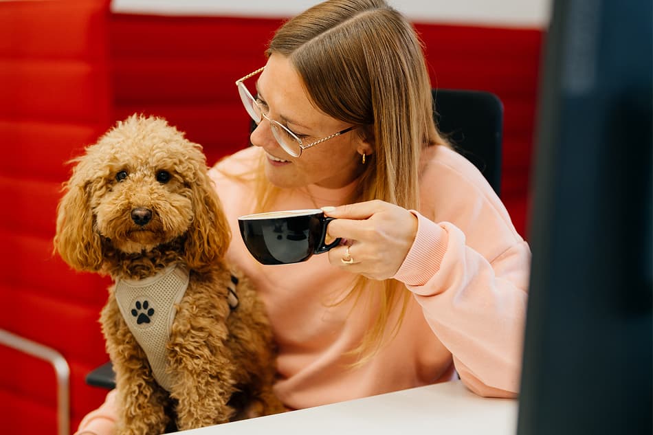 Eine Frau in einem rosa Pullover sitzt mit einem kleinen braunen Hund auf dem Schoß und hält eine schwarze Tasse in der Hand; sie befindet sich in einem Raum mit rotem Hintergrund.